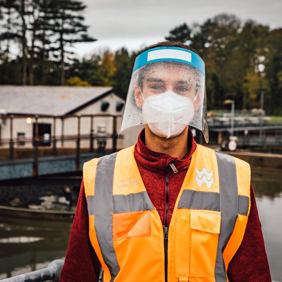 A young man standing in front of a body of water, wearing a hi-vis jacket and a facemask and visor
