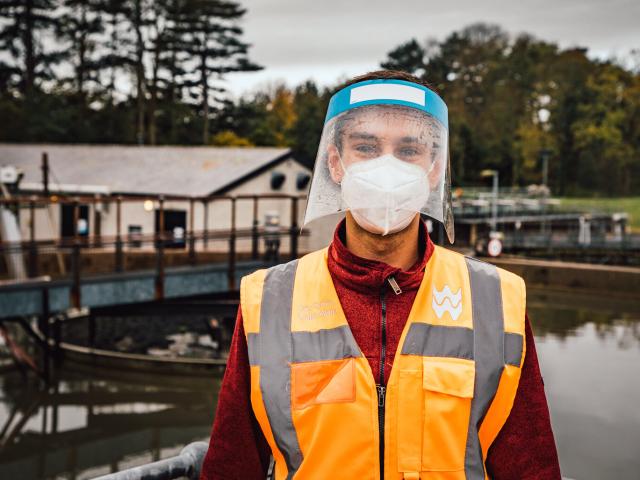 A young man standing in front of a body of water, wearing a hi-vis jacket and a facemask and visor