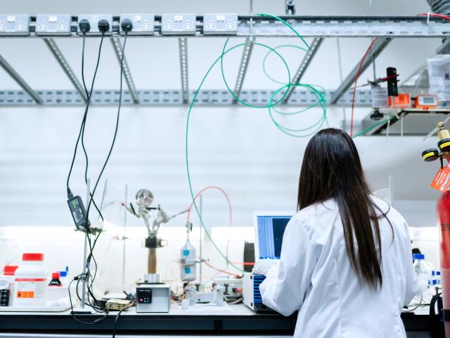 Female engineer in white coat working at a lab bench, with back to camera