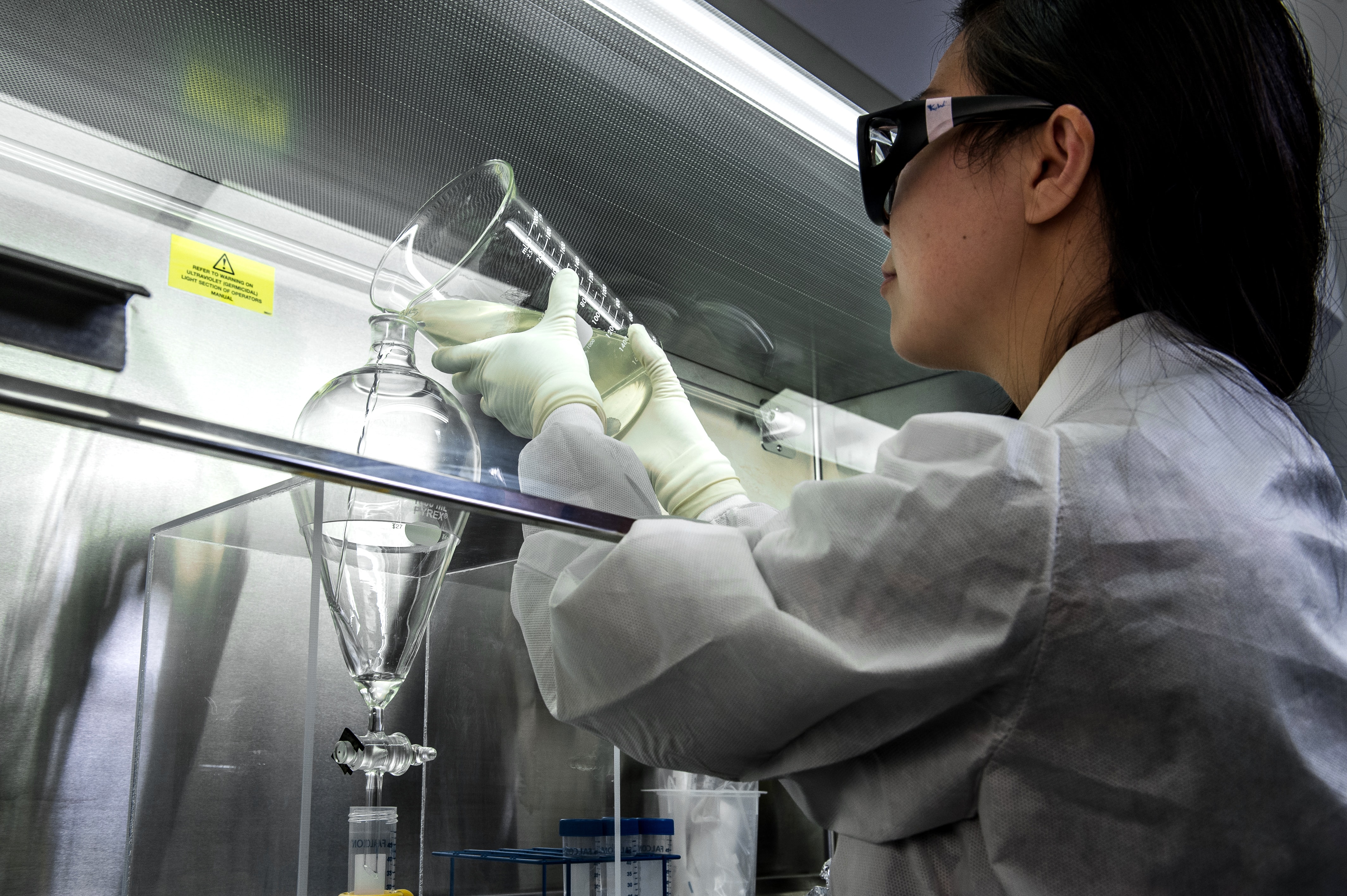 A female scientist in white lab coat and goggles pouring liquid into laboratory apparatus