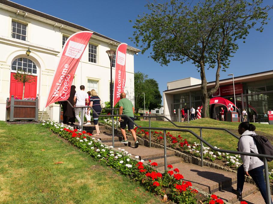 People walking up steps bordered with grass and flowers towards a white university building with red welcome flags outside