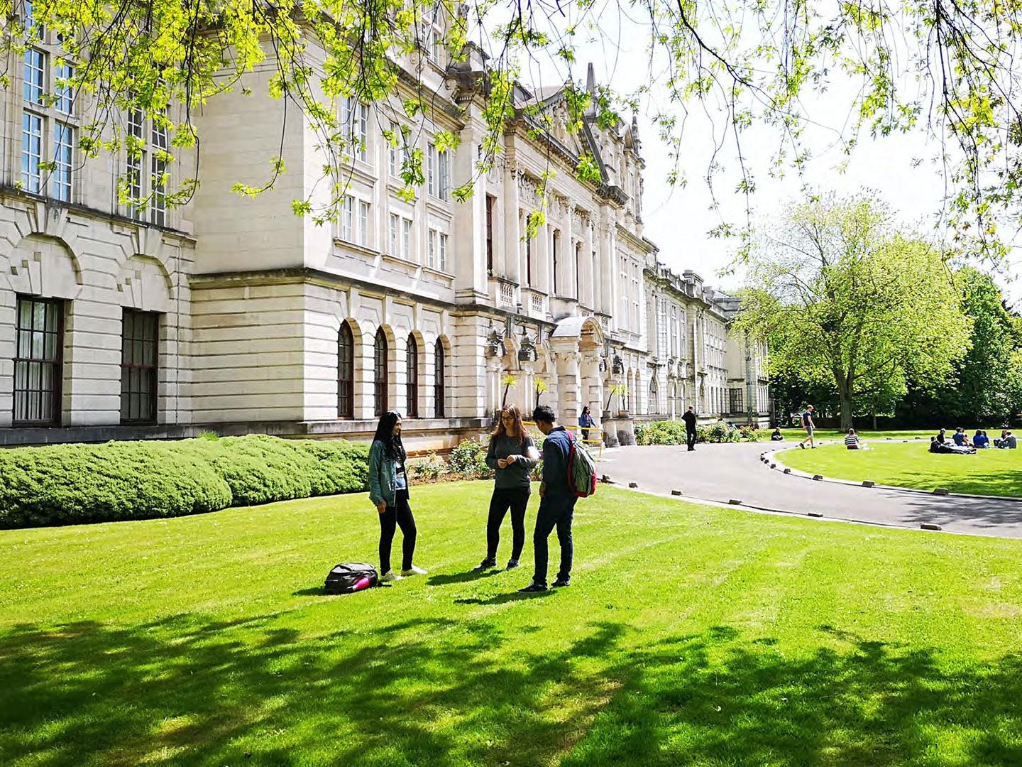 A group of student's stood outside Cardiff University Main Building, with grass, trees and other students in the background