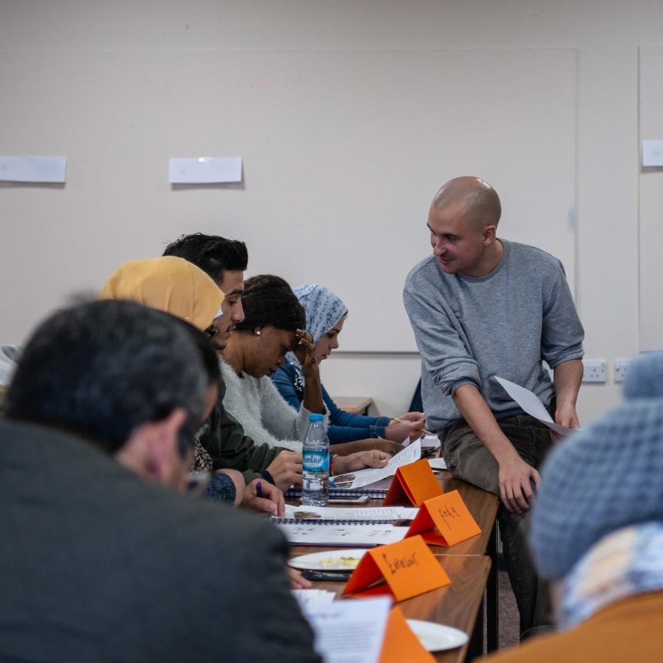 A class of adult learners with a tutor sitting on a desk