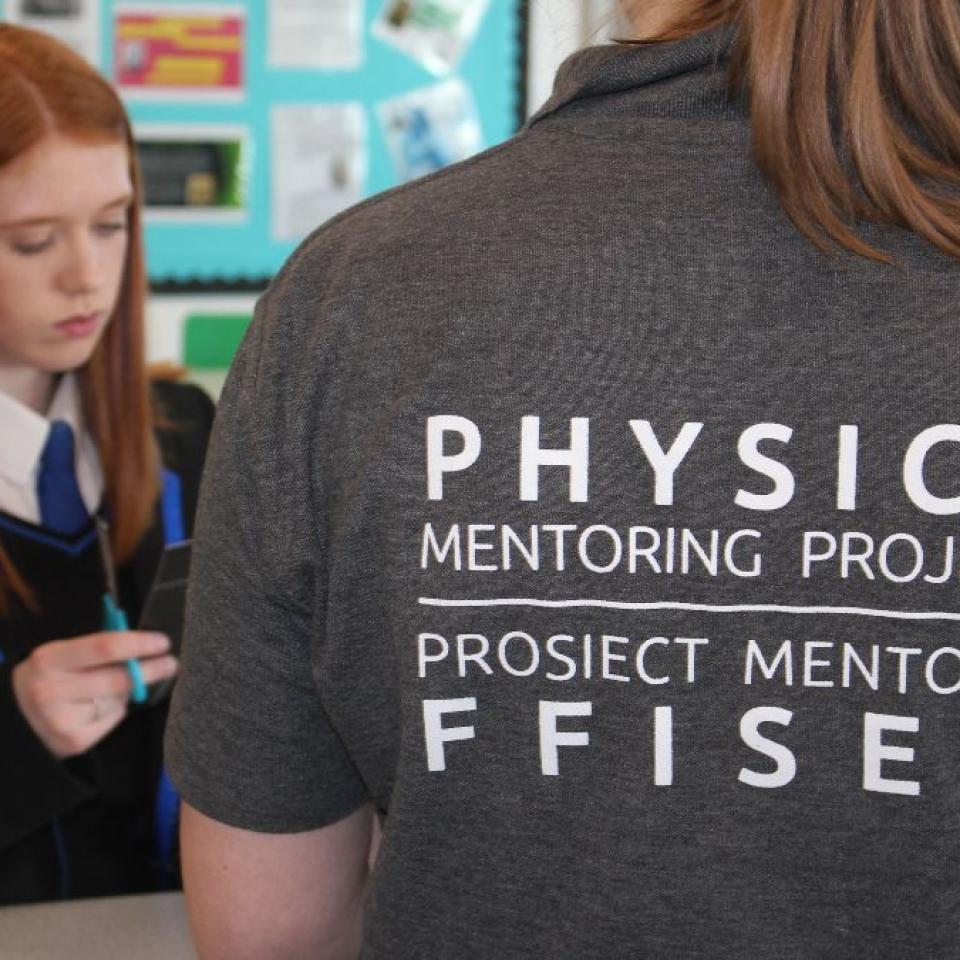 A female school student and another female with her back to the camera wearing a grey t-shirt that says Physics Mentoring Project