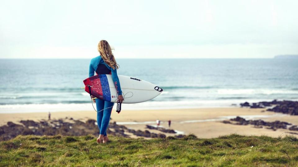 Woman on a cliff holding a surfboard and looking down on a beach