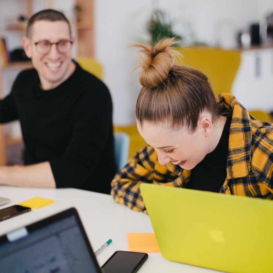 A young man and young woman working at laptops and laughing