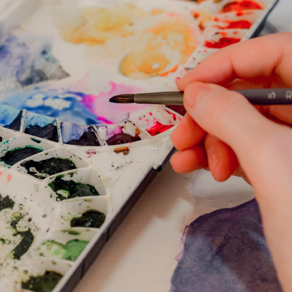 Close up shot of someone holding a paintbrush over a palette of paints