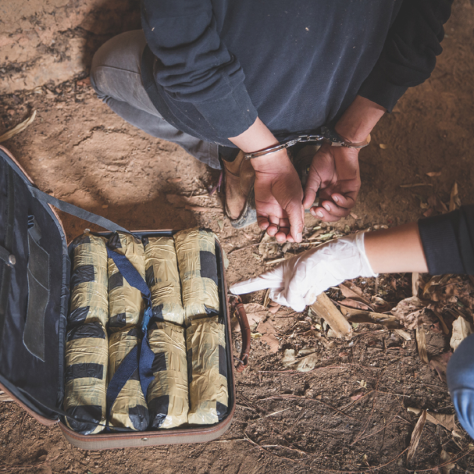 A person in handcuffs kneeling next to a bag of drugs and a person wearing latex gloves pointing at the bag