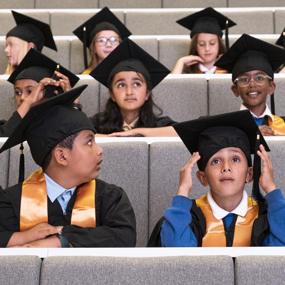 Young children in a lecture theatre wearing graduation caps and gowns