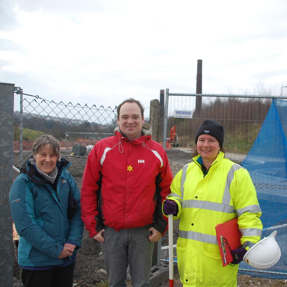 Three people standing by a chainlink fence with a sign saying 'The Hafod and Morfa Cooperworks Regeneration Project'