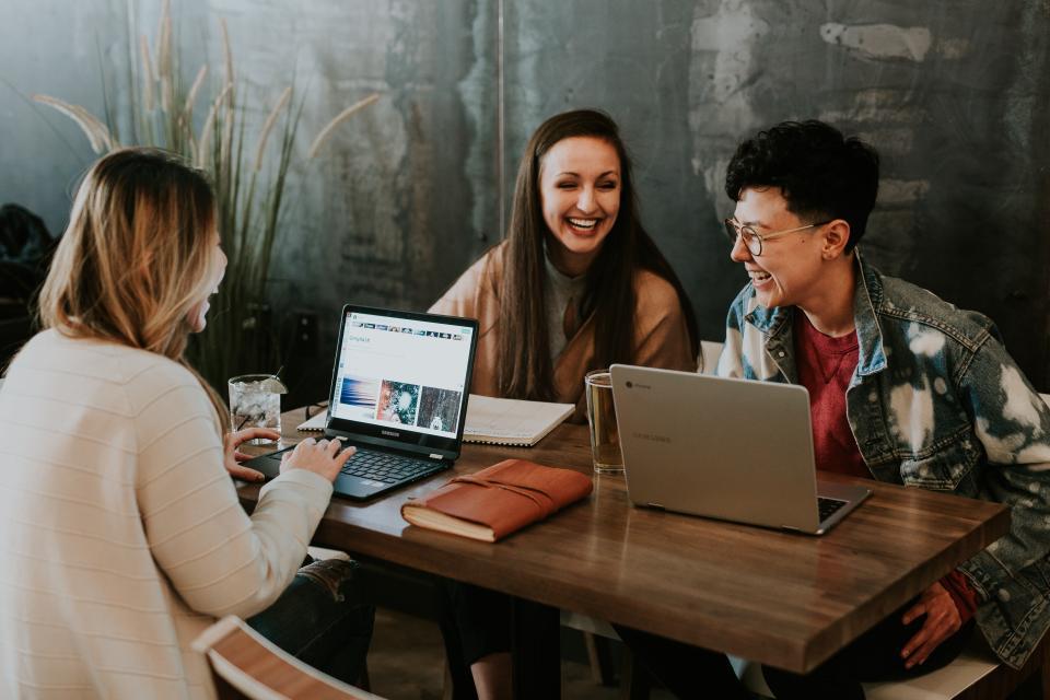 Three students sat around a table looking at laptops and smiling