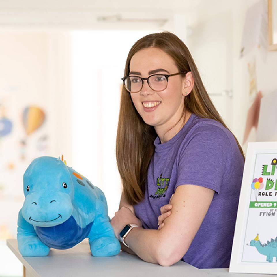 Photo of Ffion standing at a reception desk next to a blue cuddly dinosaur and a sign saying 'Little Dinos'