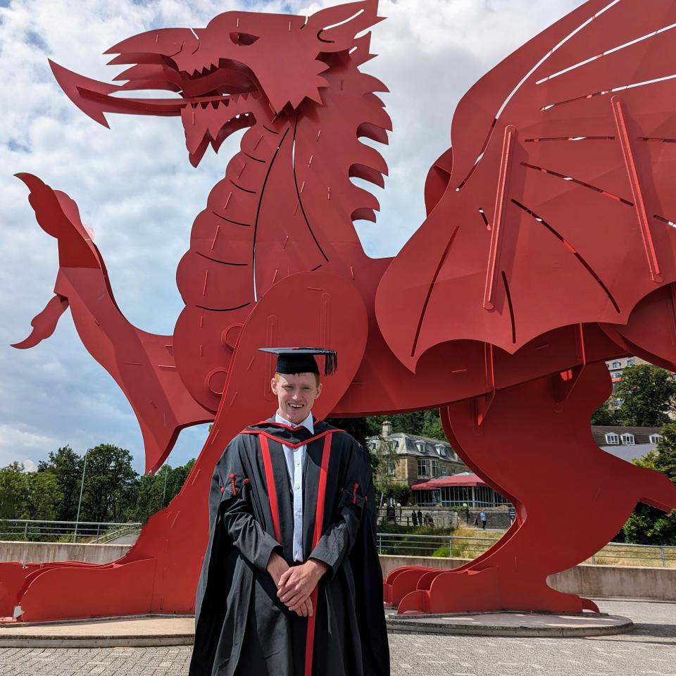 Ross in graduation cap and gown standing in front of a red dragon statue