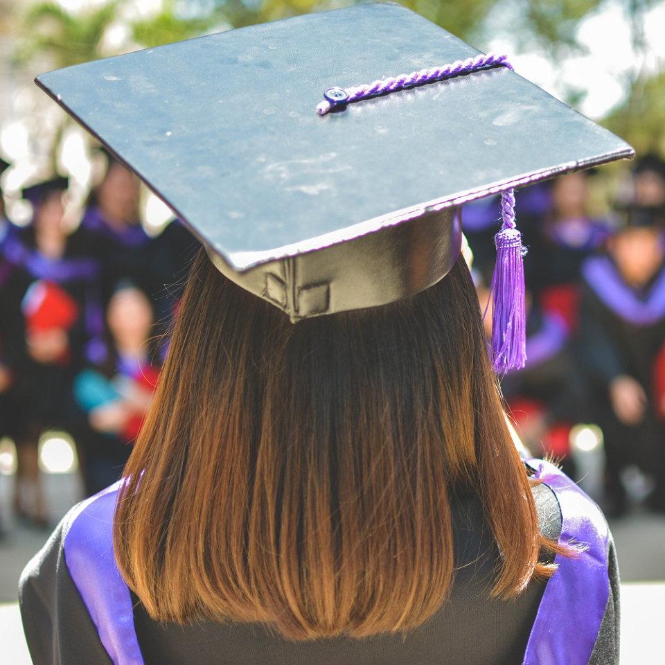 A student in graduation gowns standing in front of a seated crowd of students in graduation gowns. 