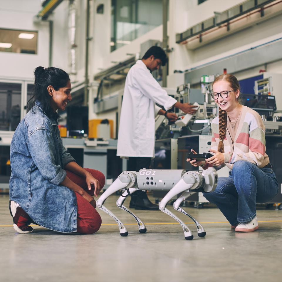 Two women working with a robot dog, with a man in a white lab coat working in the background