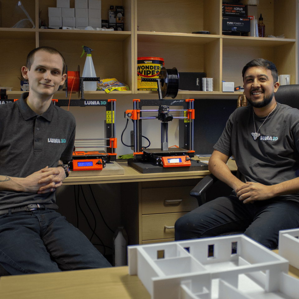 Two men sitting on chairs in an office with 3D printing equipment on the shelves behind them