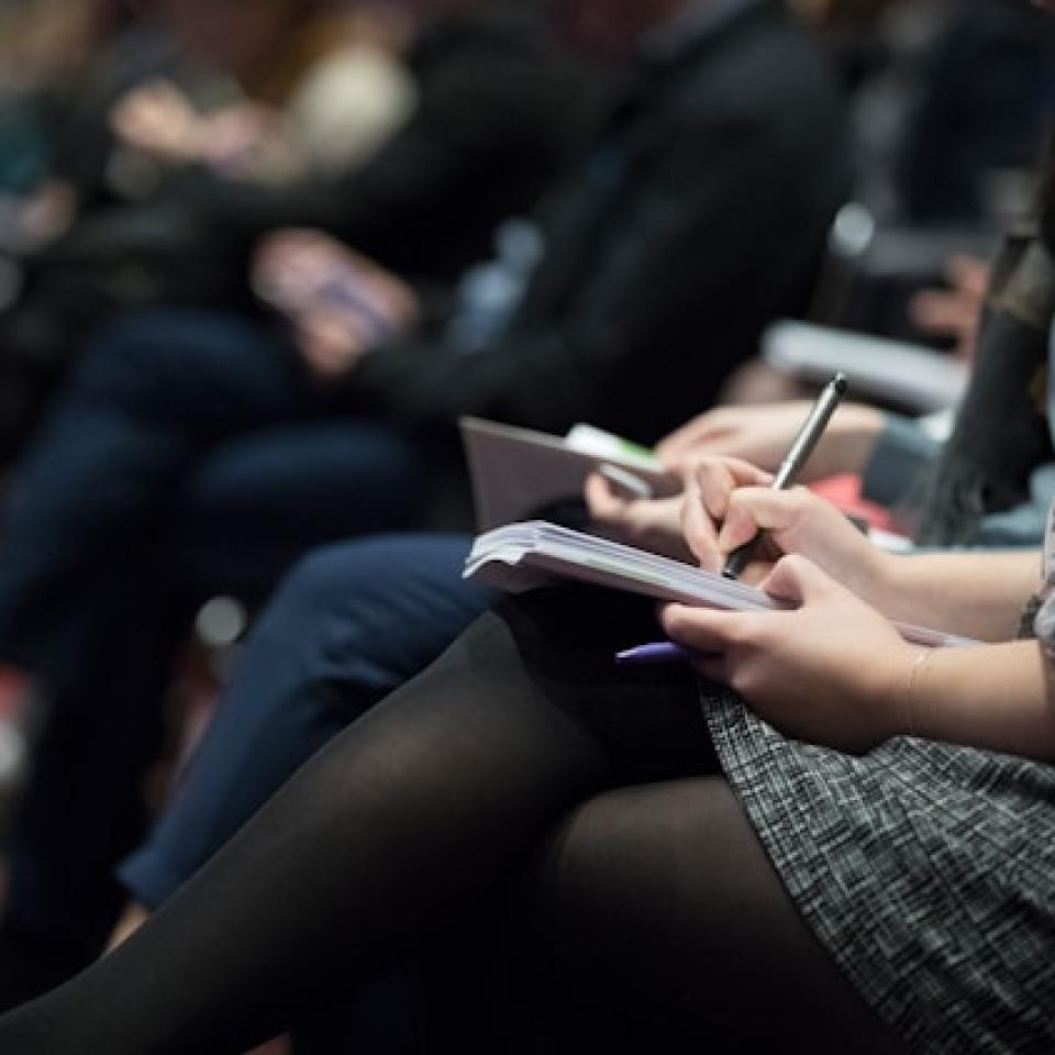 A group of people sitting in a row at a conference, taking notes