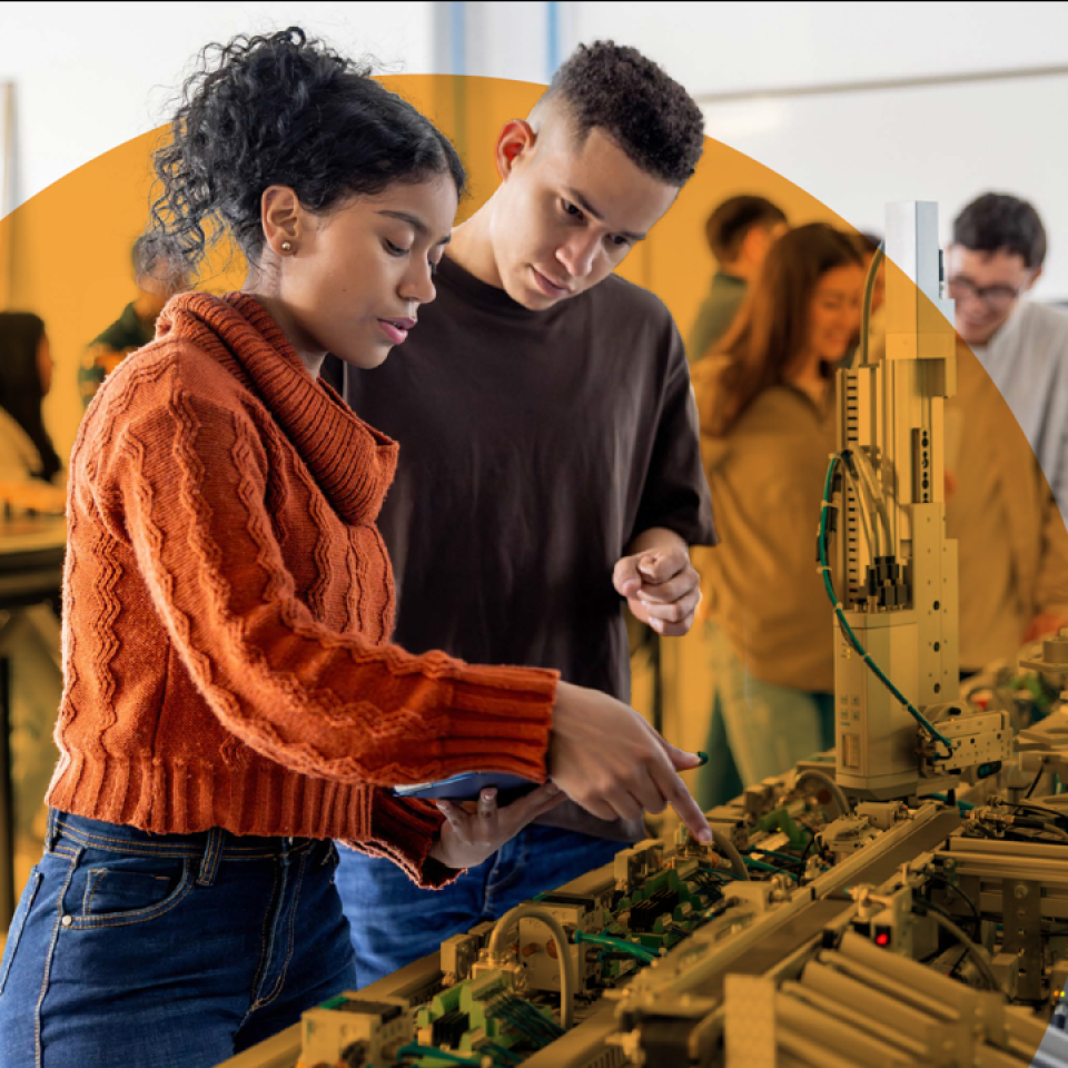 A male and female student are in a large room looking at equipment. Other students are in the background