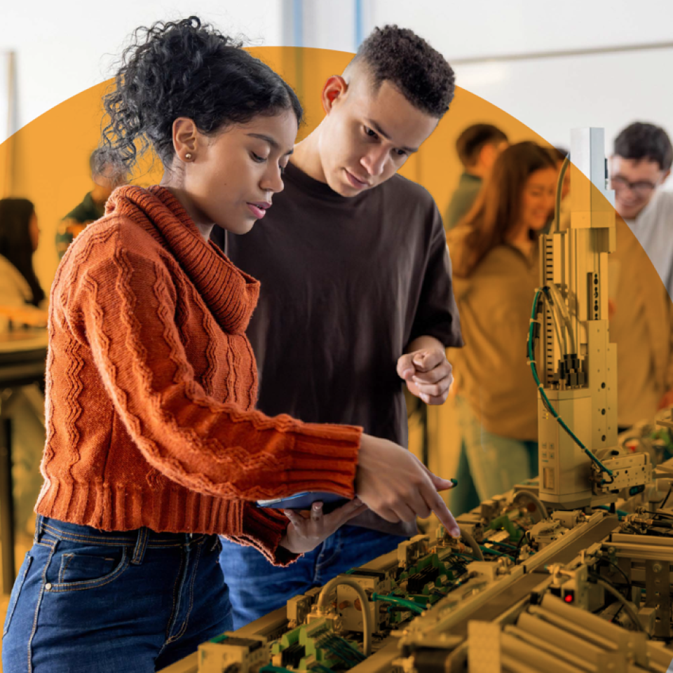A male and female student are in a large room looking at equipment. Other students are in the background
