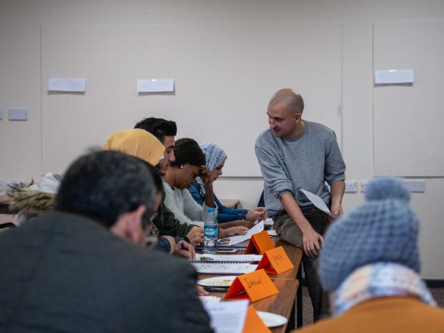 A class of adult learners with a tutor sitting on a desk