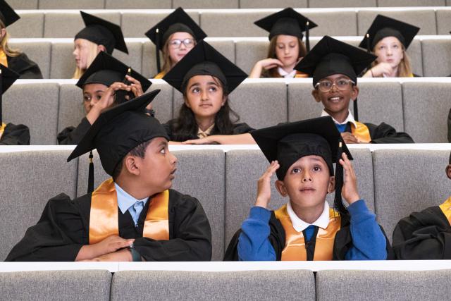 A group of children in graduation caps and gowns sitting in a lecture theatre