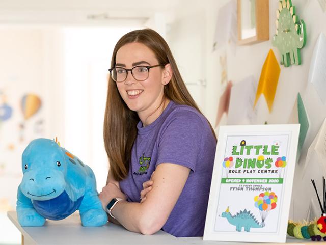 Photo of Ffion standing at a reception desk next to a blue cuddly dinosaur and a sign saying 'Little Dinos'
