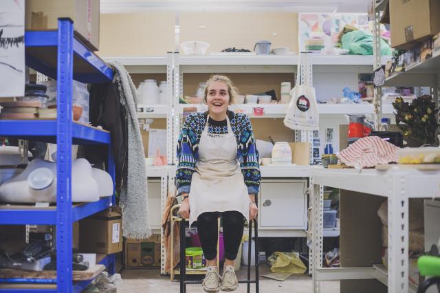 A photo of a smiling woman in an apron sitting on a stool in a workroom