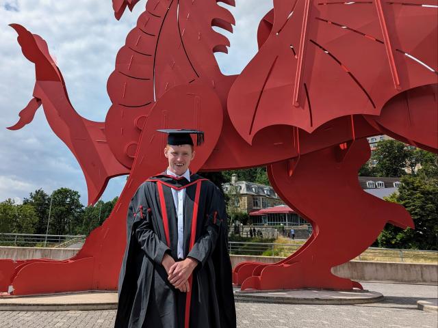 Ross in graduation cap and gown standing in front of a red dragon statue