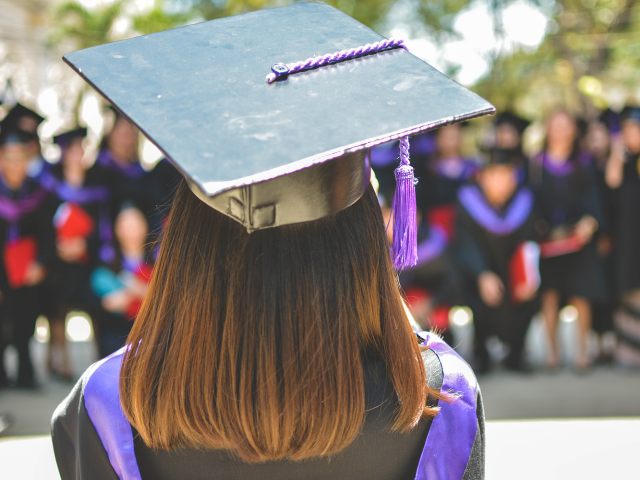 A student in graduation gowns standing in front of a seated crowd of students in graduation gowns. 