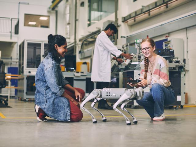 Two women working with a robot dog, with a man in a white lab coat working in the background