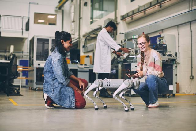 Two women working with a robot dog, with a man in a white lab coat working in the background
