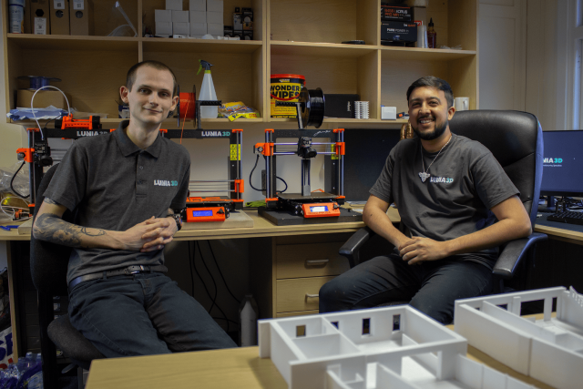 Two men sitting on chairs in an office with 3D printing equipment on the shelves behind them