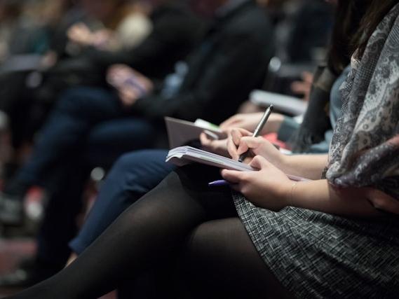 A group of people sitting in a row at a conference, taking notes
