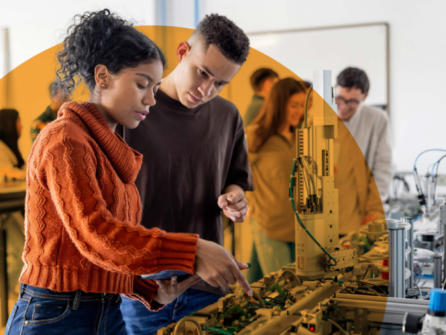 A male and female student are in a large room looking at equipment. Other students are in the background